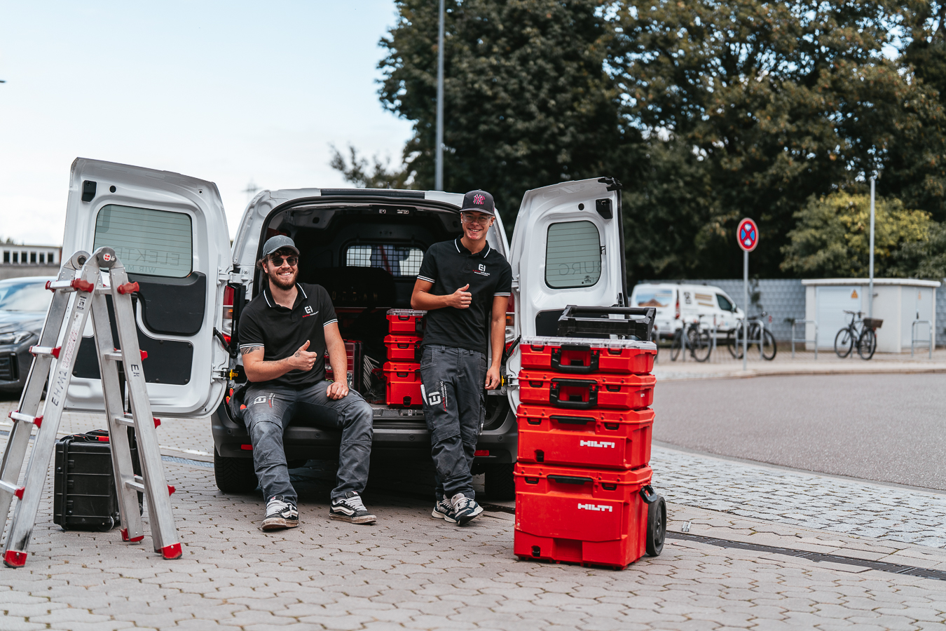 Zwei laechelnde Handwerker in schwarzen Shirts am Transporter mit roten Werkzeugkoffern